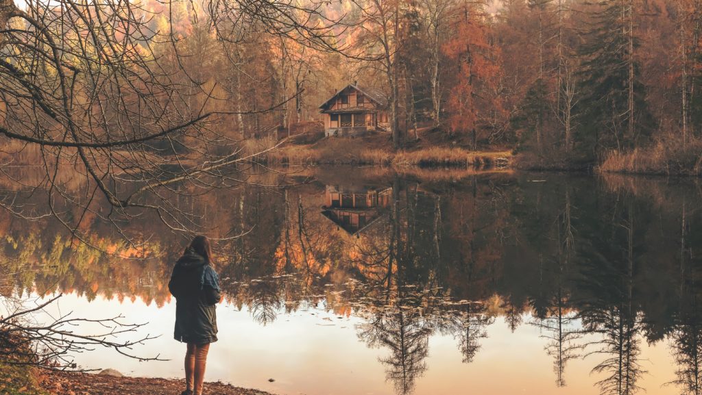 A woman regarding a lake with a cabin among trees in autumn