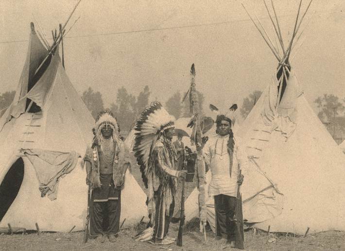 An old photograph of Native Americans with headdresses of feathers and wigwams behind them