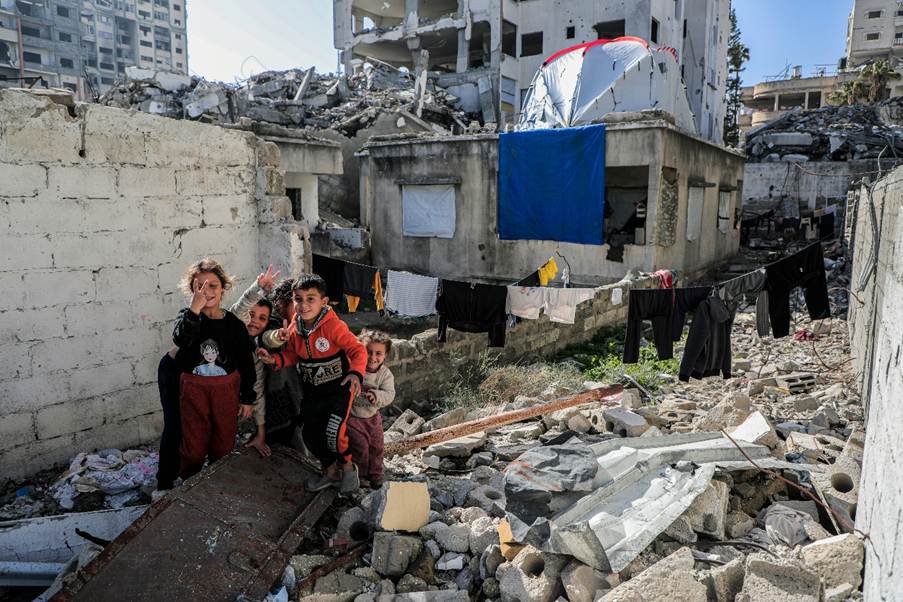 Some children smiling cheerfully beside bombed-out buildings in Gaza